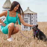 Woman in teal dress sitting with a dog in a field with patterned huts in the background